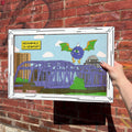 Hand holding a framed drawing of the purple people eater bridge and purple people eater against a brick wall.